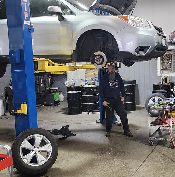 A mechanic stands beneath the car, holding a tool and car being lifted to get serviced.